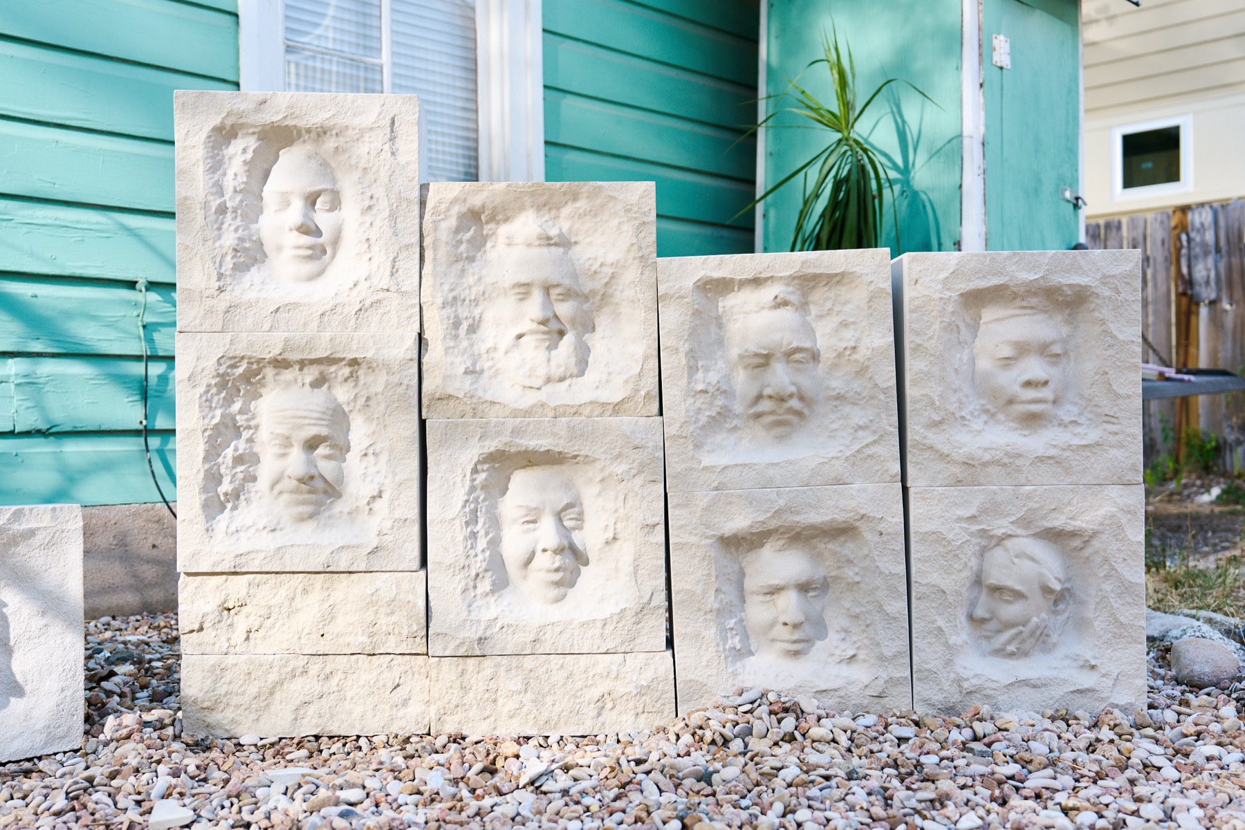 Eight limestone blocks, each carved with a different face, stacked on gravel in front of a teal house.